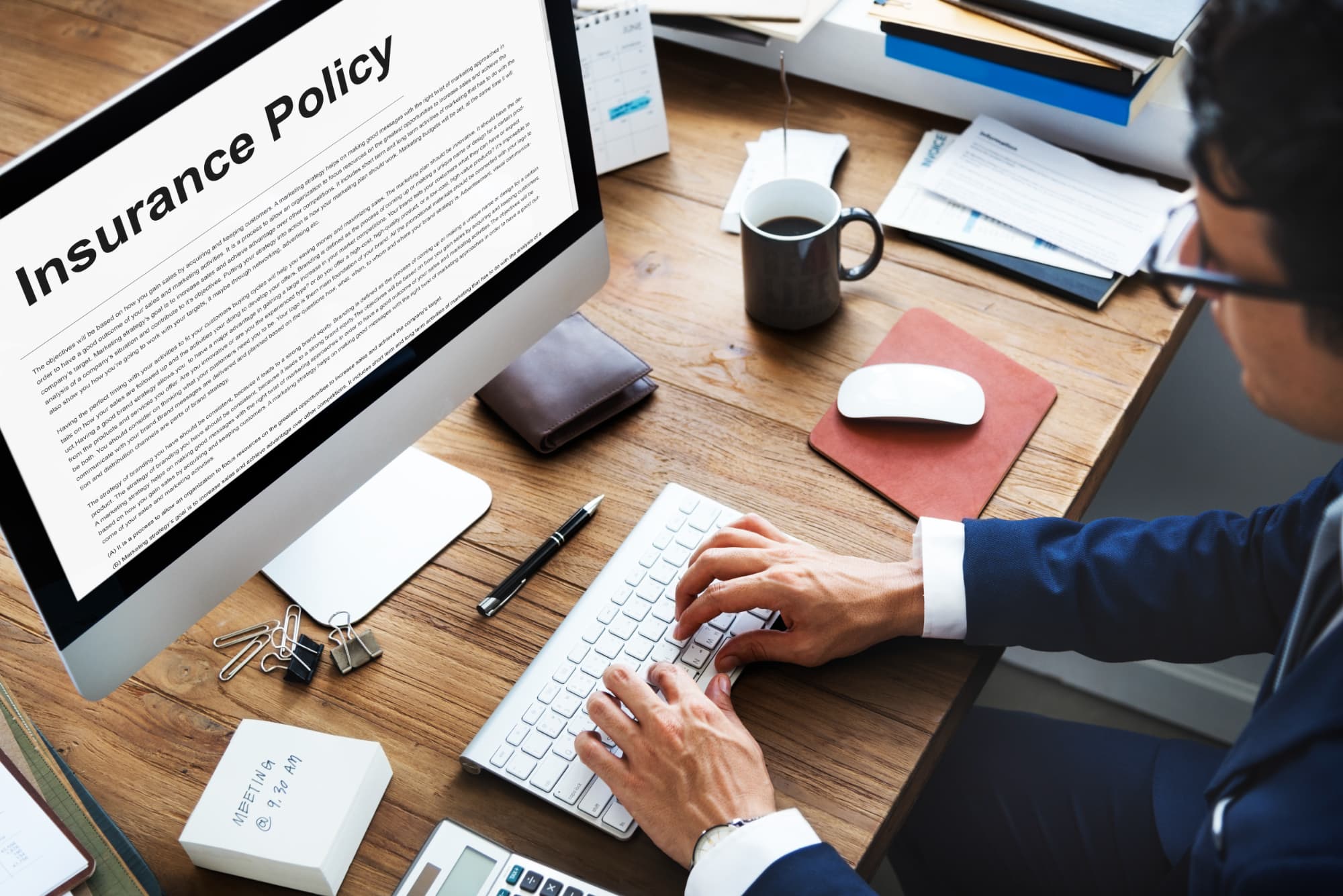 A person in business attire types on a keyboard at a desk, reviewing an insurance policy document with "Illnesses Covered By TPD" displayed on a large computer monitor. Office supplies, a phone, and a wallet are also on the wooden desk.