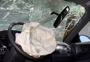 The interior of a car with a deployed airbag, shattered windshield, and broken rearview mirror suggests a recent collision or accident with potential vehicle injuries.