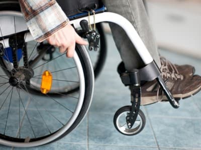 A close-up of a person with total and permanent disability in a wheelchair, wearing brown shoes and gray pants, with one hand on the wheel and the other adjusting the wheel lock on a tiled floor.