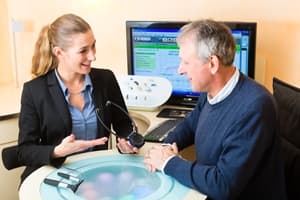 A woman in business attire shows a hearing aid device to an older man seated at a desk, discussing ways to prevent medical negligence, while a computer screen displaying data glows in the background. Both appear engaged in conversation.