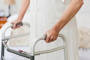 An injured worker wearing a white robe is standing and holding onto a walker for support in a bedroom. Only their hands and part of their body are visible.