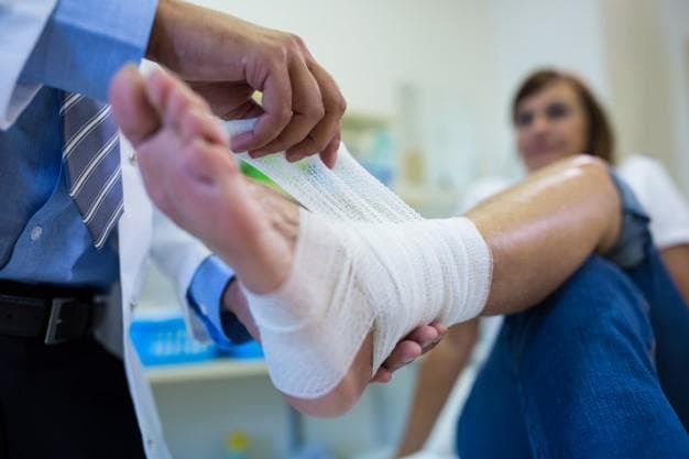 A medical professional wraps a bandage around a patient's injured foot while the patient sits on an exam table in a clinic.