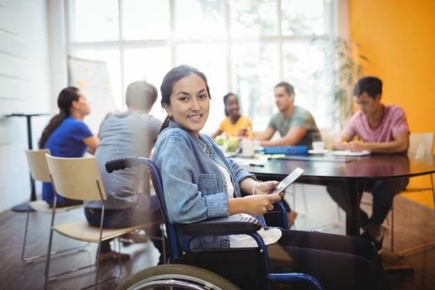 A woman in a wheelchair smiles at the camera while holding a tablet. She is in a meeting room with a group of people engaged in discussion around a table in the background.