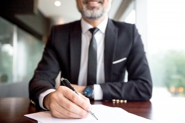 A man in a suit sitting at a desk, smiling, and holding a pen while writing on a sheet of paper.