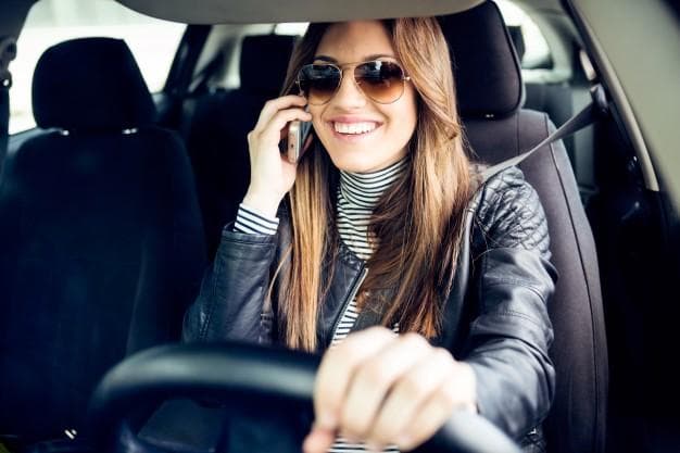 A smiling woman wearing sunglasses and a leather jacket is talking on her phone while driving a car. She has one hand on the steering wheel and appears cheerful.