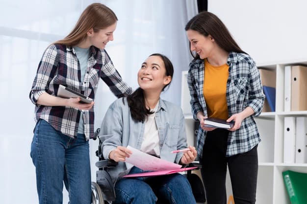 Three young women, one in a wheelchair holding papers, smile and talk together indoors. Two others stand beside her, holding books and a tablet, showing a supportive and friendly atmosphere.