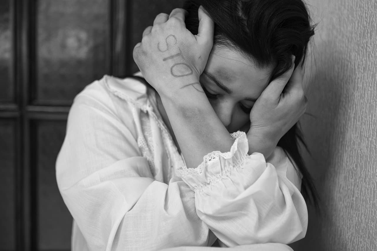 A distressed woman sits against a wall, covering her face with her hands. The word "STOP" is written on her forearm, and she has visible bruises, suggesting she is a victim of abuse.