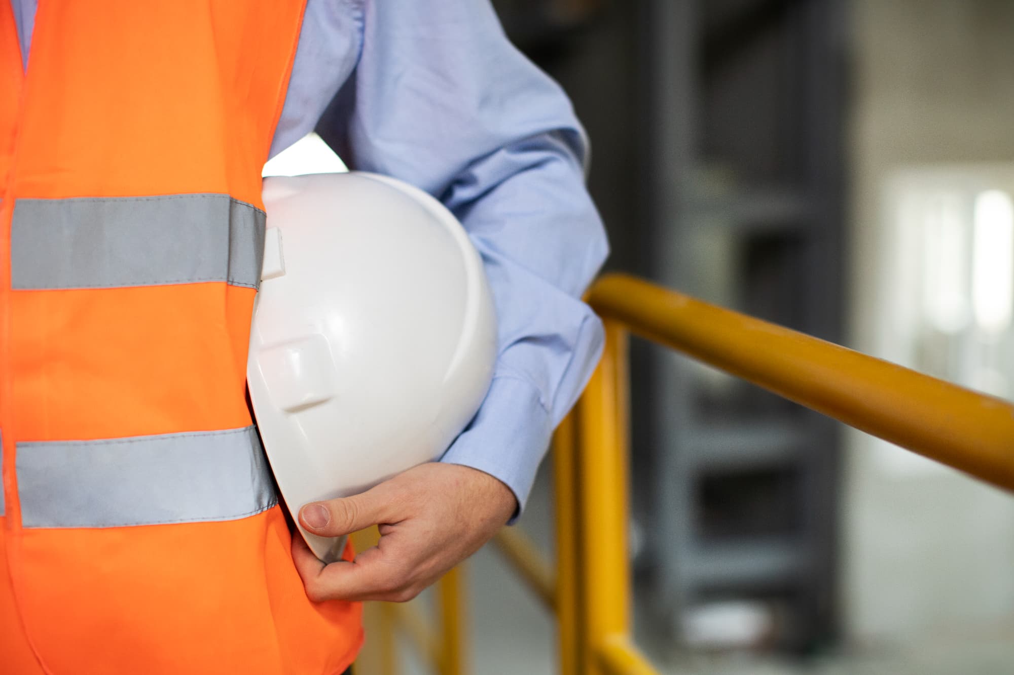A person wearing a blue shirt and orange reflective safety vest holds a white hard hat while standing next to a yellow railing, highlighting the importance of safety to prevent construction site injuries in an industrial setting.