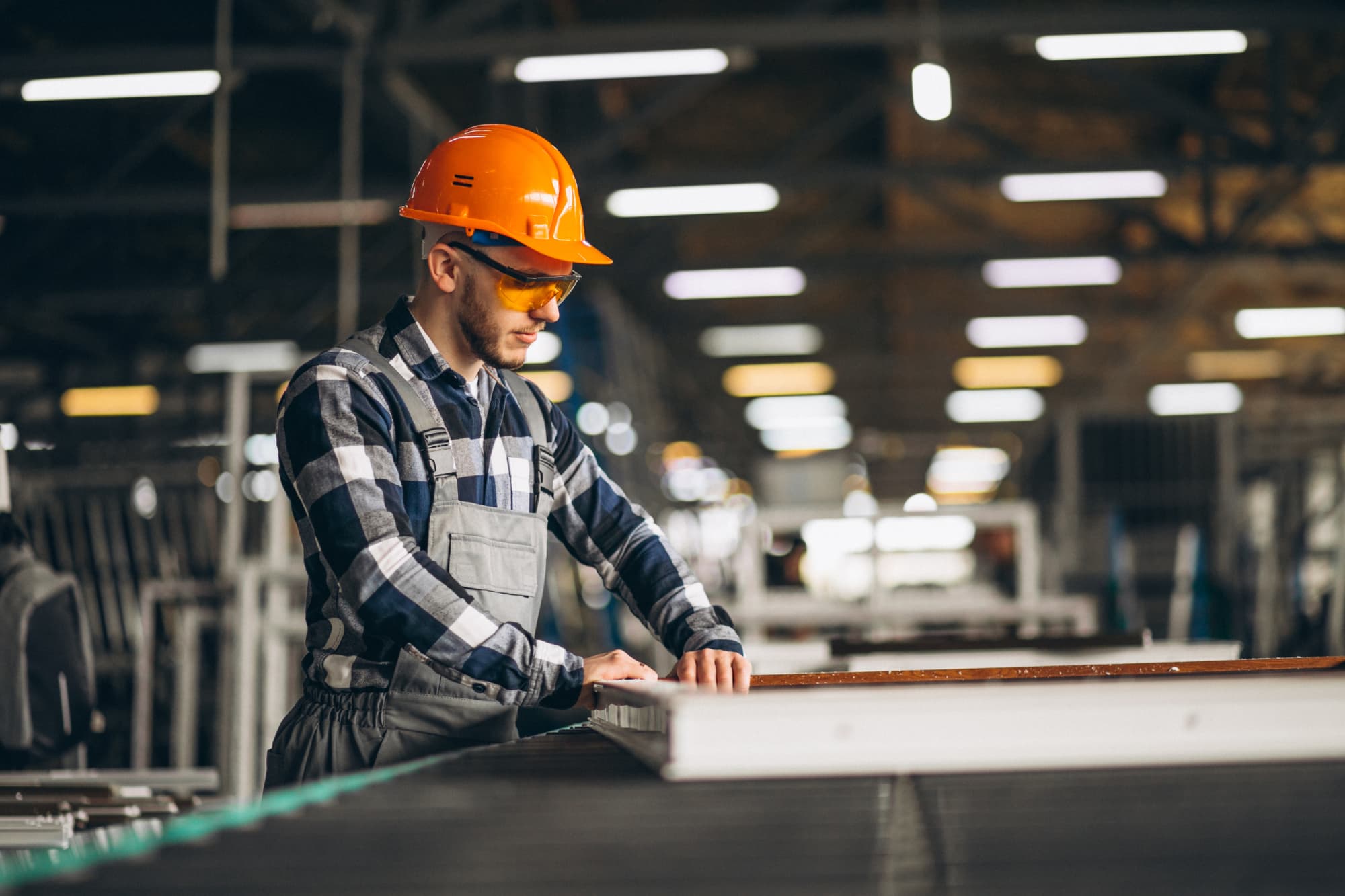 A factory worker wearing a plaid shirt, overalls, an orange hard hat, and safety glasses inspects a panel on a production line in a brightly lit industrial setting, maintaining safety to help prevent factory worker injuries.