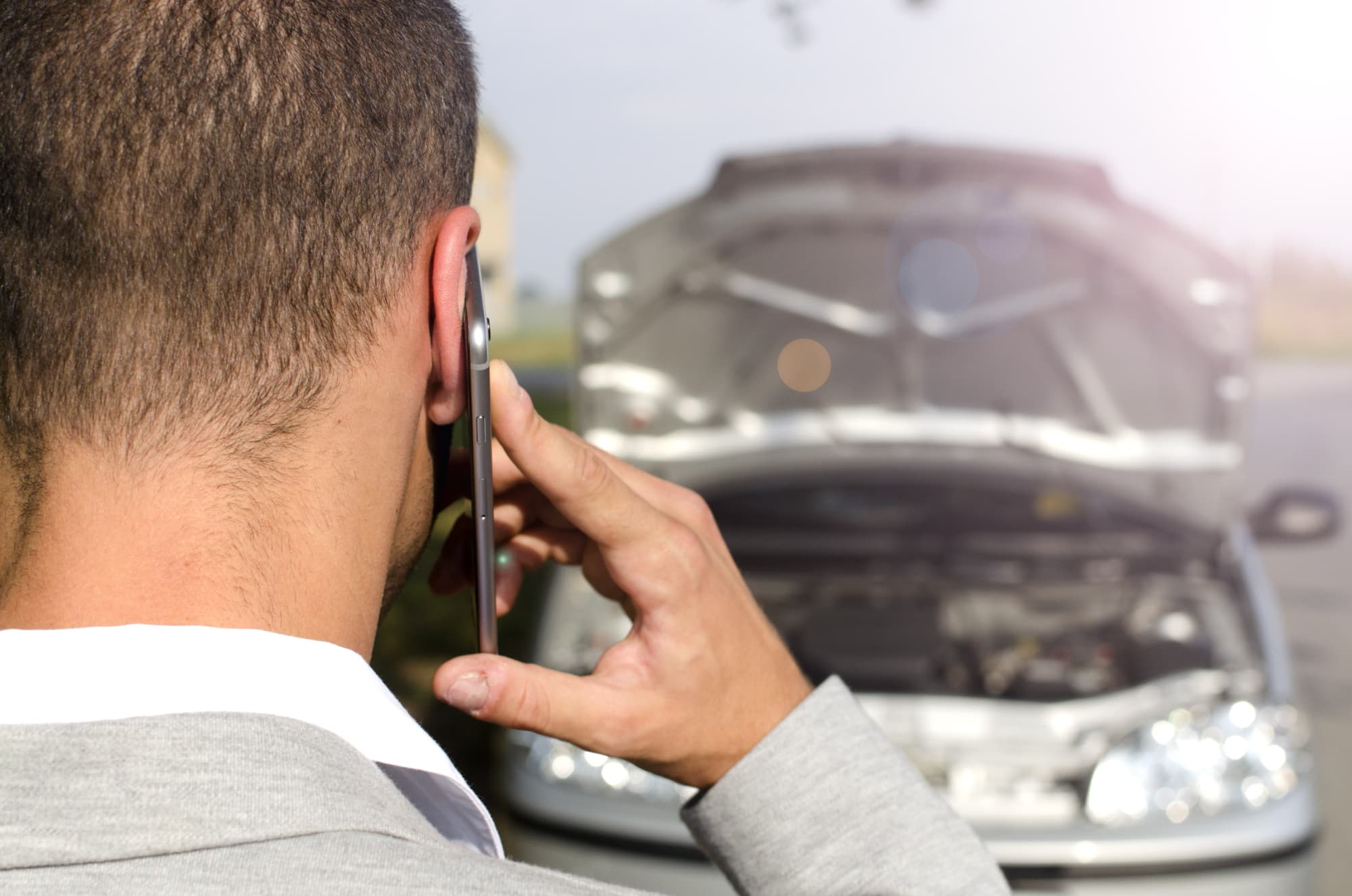 A man in a suit is seen from behind, holding a phone to his ear, standing before a car with its hood open—suggesting a possible breakdown or seeking Car Accident Injury Compensation NSW.