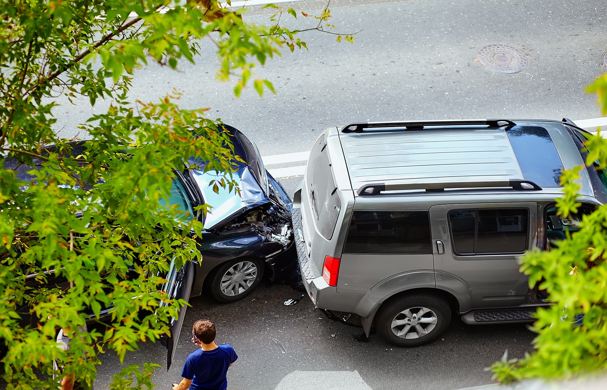 Aerial view of a traffic accident shows a black car crashed into the back of a gray SUV on a city street, with a person standing nearby and green tree branches framing the image.