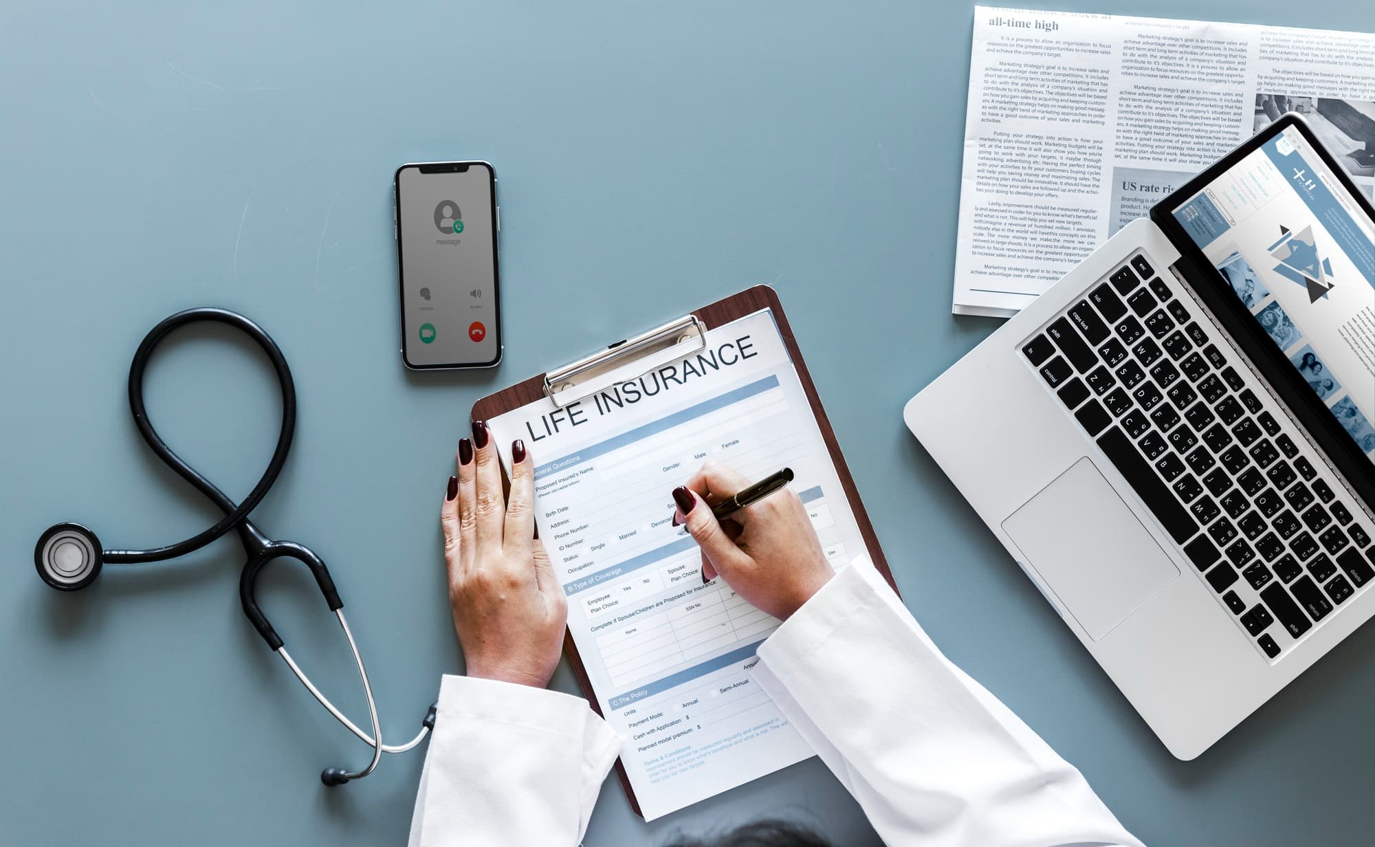 A person in a white coat fills out a life insurance form on a clipboard, with documents about head trauma, a laptop, stethoscope, and a smartphone with an incoming call on a gray desk.