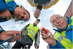 Four emergency responders in reflective vests and jackets look down toward the camera, one holding a stethoscope and another a bloody bandage, suggesting a medical emergency scene—a situation where public liability lawyers may be needed. The sky is visible in the background.