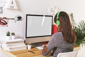 A person with long hair wearing green headphones sits at a desk, working on a computer with a blank screen—perhaps researching Total and Permanent Disability. The desk has stacked papers, cacti, and a lamp, with plants and wall decor in the background.