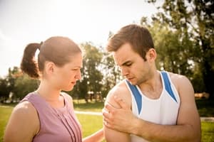 A woman comforts a man outdoors as he holds his shoulder in pain, possibly considering injury compensation. They appear to be in a park with trees and sunlight in the background.