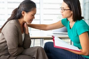 A woman in a brown jacket looks upset while sitting next to another woman in glasses and a teal shirt, who is comforting her about injury compensation with a hand on her shoulder and holding a folder.