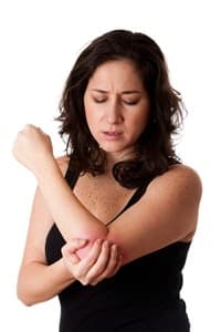Woman with medium-length brown hair wearing a sleeveless black top, holding and rubbing her left elbow with a pained expression, suggesting discomfort or injury possibly due to a recent motor accident.