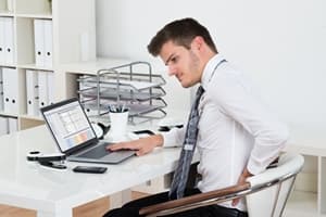A man in business attire sits at a desk using a laptop, his strained expression and hand on his back suggesting discomfort or back pain—an issue often addressed through workers compensation. Office supplies and shelves fill the background.