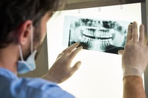 A person wearing gloves and a mask examines a dental X-ray image on a lightbox, carefully checking for signs that could indicate dental malpractice in the clear view of teeth and jaw.