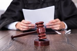 A judge in a black robe sits at a wooden desk, holding papers—perhaps reviewing if a Will can be challenged—with a wooden gavel and sound block in the foreground.