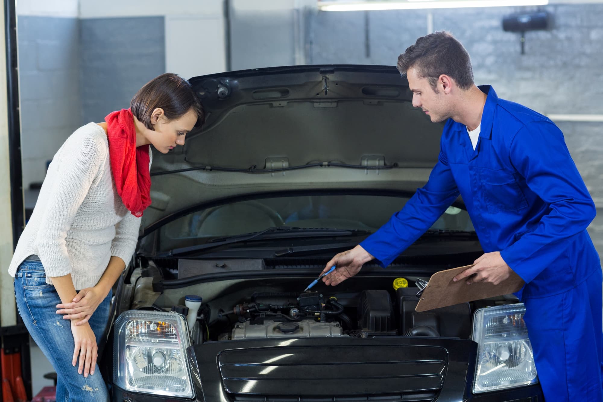 A mechanic in blue coveralls explains something under the open hood of a car to a woman in a white sweater and red scarf, possibly after a car accident Sydney locals might face. The woman leans forward attentively in the garage setting.