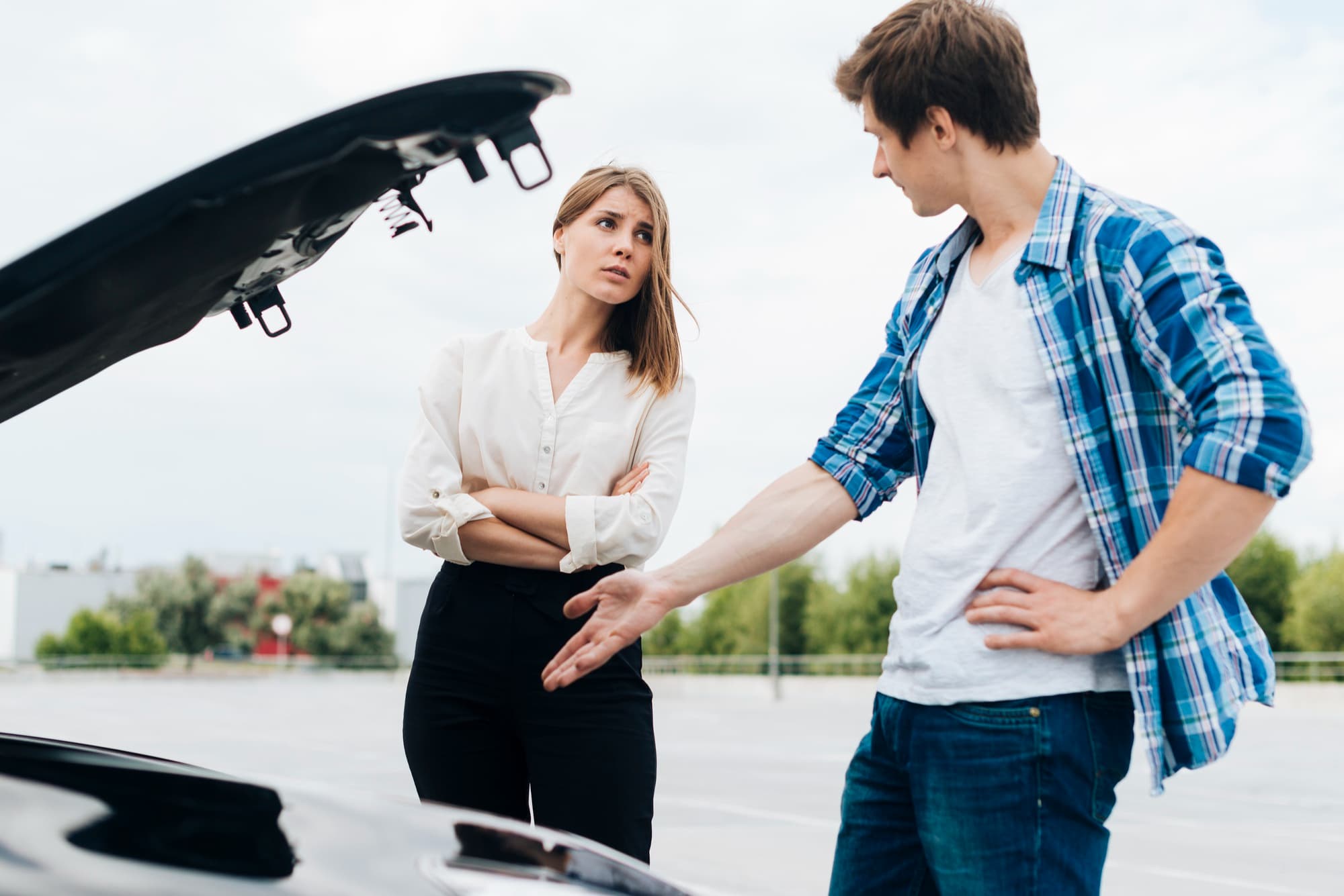 A woman stands with arms crossed beside a car with its hood open, looking at a man who gestures towards the car. They appear to be discussing motor accidents in an outdoor parking lot.