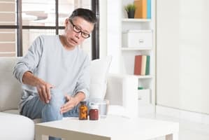 An older man sits on a couch, touching his knee in discomfort. Pill bottles and a glass of water on the table suggest he may be considering medication for pain relief after suffering private property injuries.