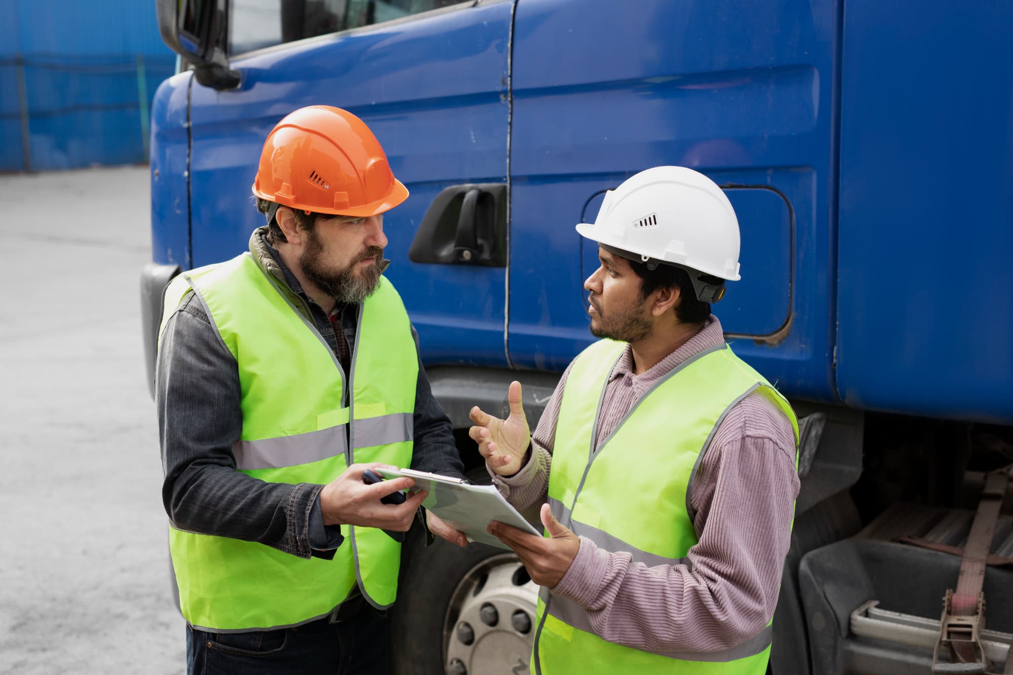 Two workers wearing safety vests and helmets stand next to a blue truck, engaged in discussion about Workers Compensation. One holds a tablet while the other holds a pen, suggesting an inspection or work-related review.