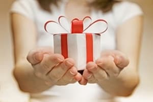 A person in a white shirt holds out a small round gift box wrapped with a red ribbon and bow, offering it with both hands—a gentle gesture contrasting the seriousness of topics like medical negligence. The background is softly blurred.