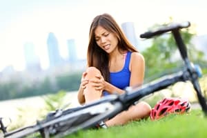 A woman sits on grass next to a fallen bicycle, holding her knee in pain, with a red helmet nearby and a blurred cityscape behind—prompting the question: Are cycling accidents increasing?.