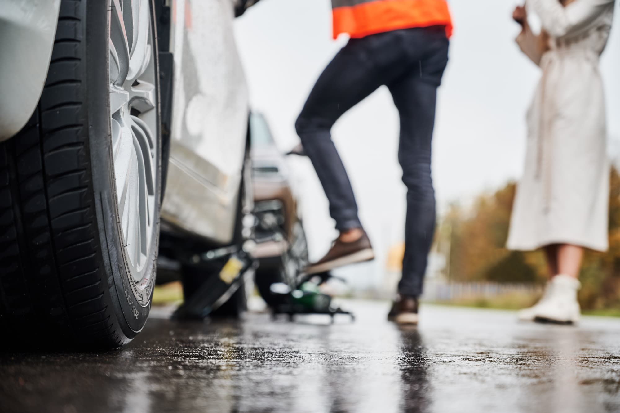 Close-up of a car tire being changed on a wet road after a car accident; one person in a reflective vest works near the car while another stands nearby in a light-colored coat.