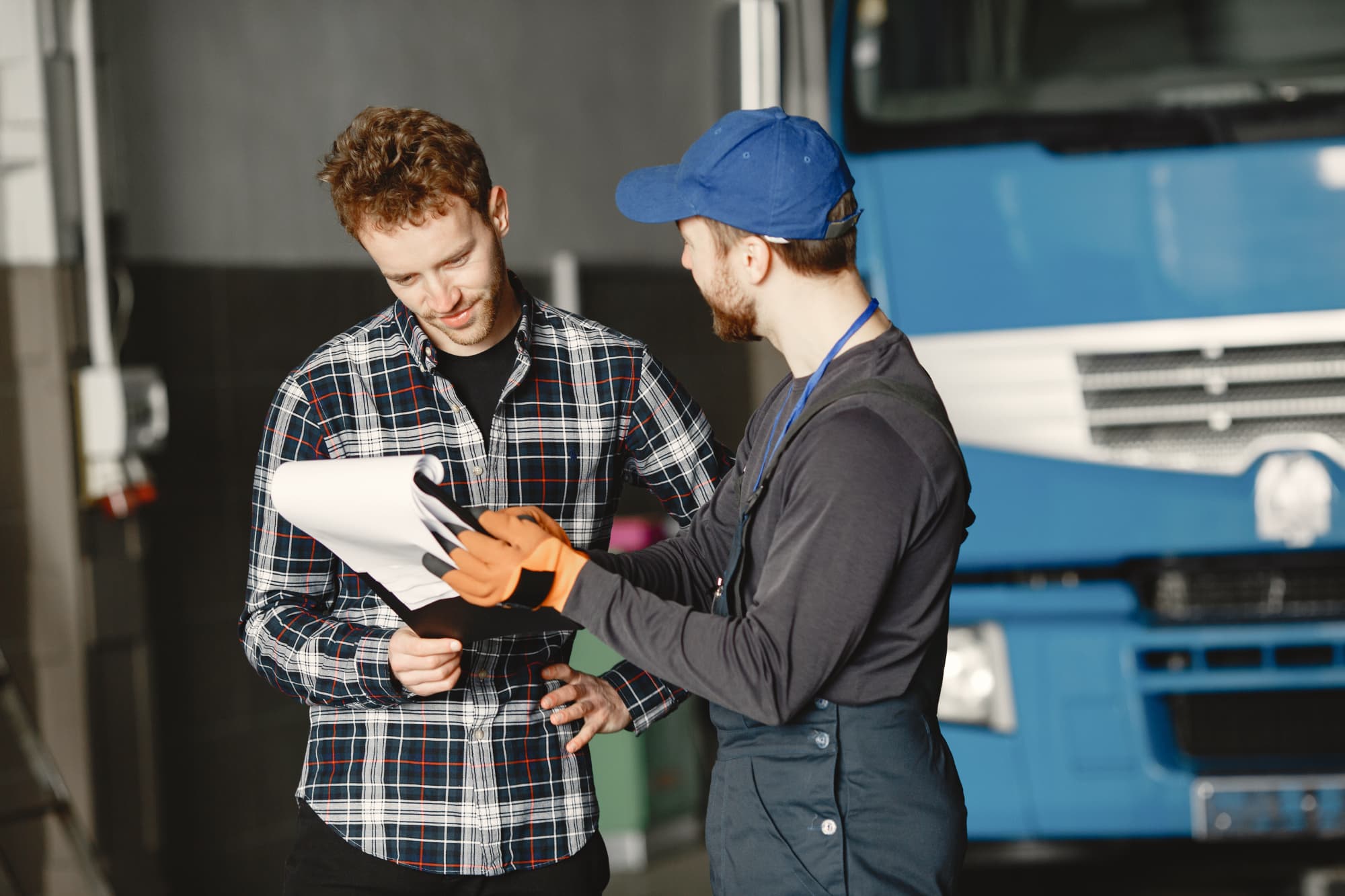 A mechanic wearing gloves and a cap shows a clipboard to a man in a plaid shirt as they discuss paperwork about accidents involving trucks, standing in front of a large blue truck inside the garage.
