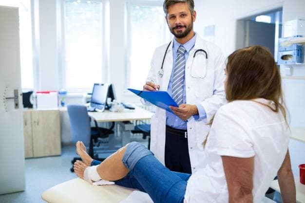 A doctor with a stethoscope and clipboard talks to a patient with a bandaged knee, who is sitting on an examination table in a medical office.