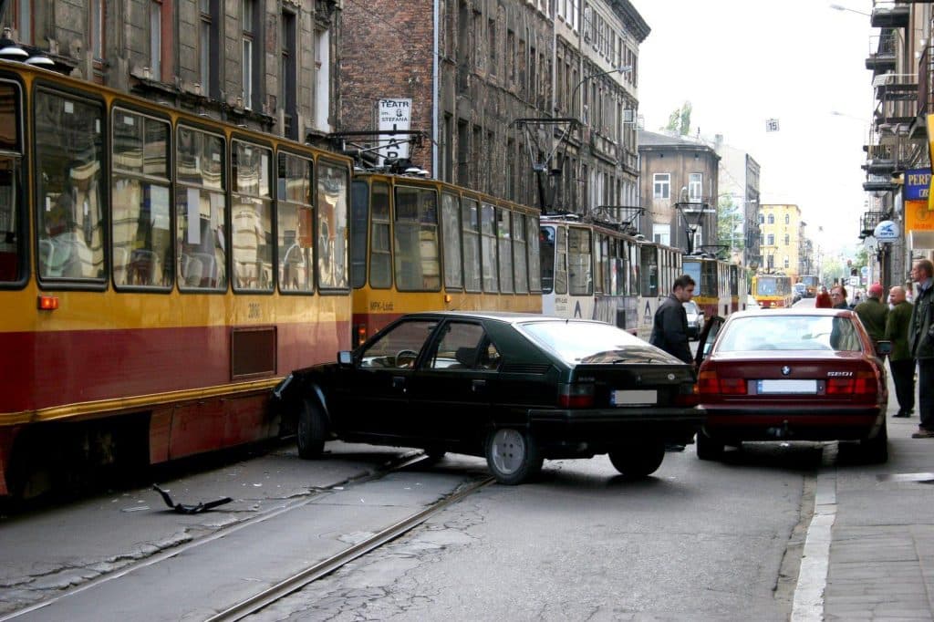 A black car has crashed into a red and yellow tram in a city street, partially blocking tram tracks. Another red car is parked nearby, and several people are standing on the sidewalk observing the scene.