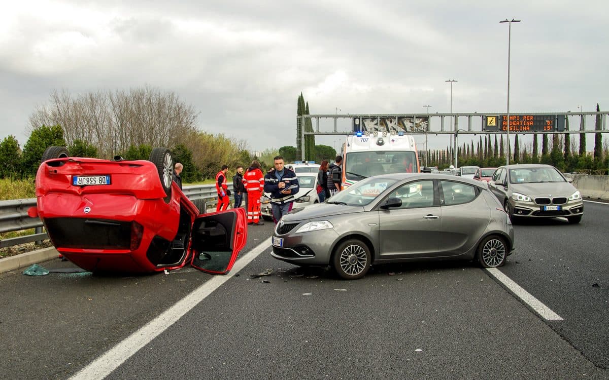 A red car is overturned on a highway next to a silver car with front damage. Emergency responders, an ambulance, and several people are at the scene. Debris is scattered on the road under cloudy skies.