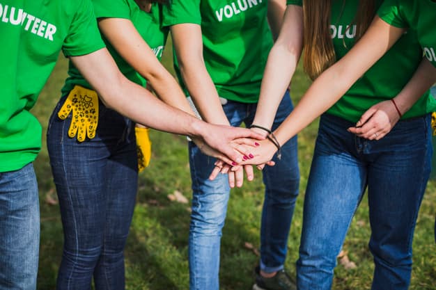 Five people wearing green "Volunteer" shirts stand in a circle outdoors and stack their hands together in the center, symbolizing teamwork and unity. The grass is visible underfoot.