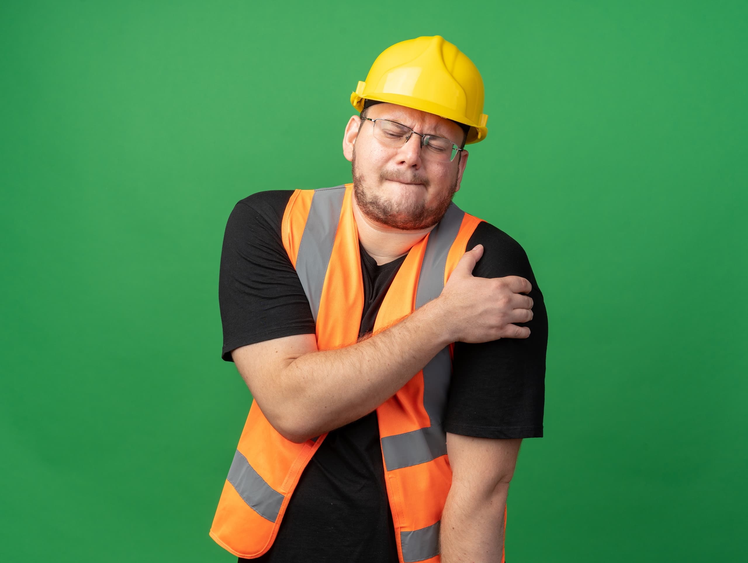 builder man in construction vest and safety helmet looking unwell touching his shoulder feeling pain standing over green background