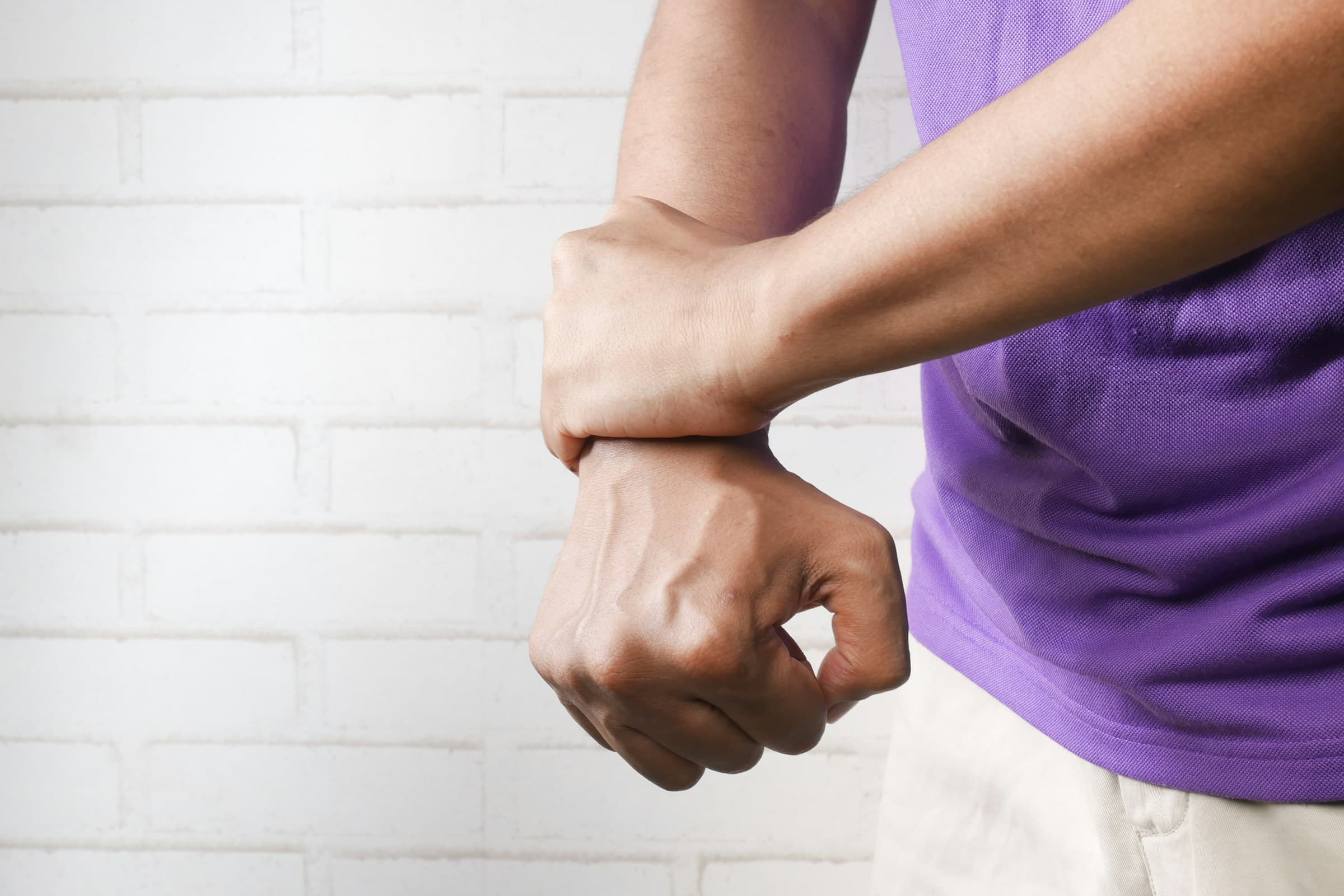 A person wearing a purple shirt and light pants holds their right wrist with their left hand against a white brick wall background.